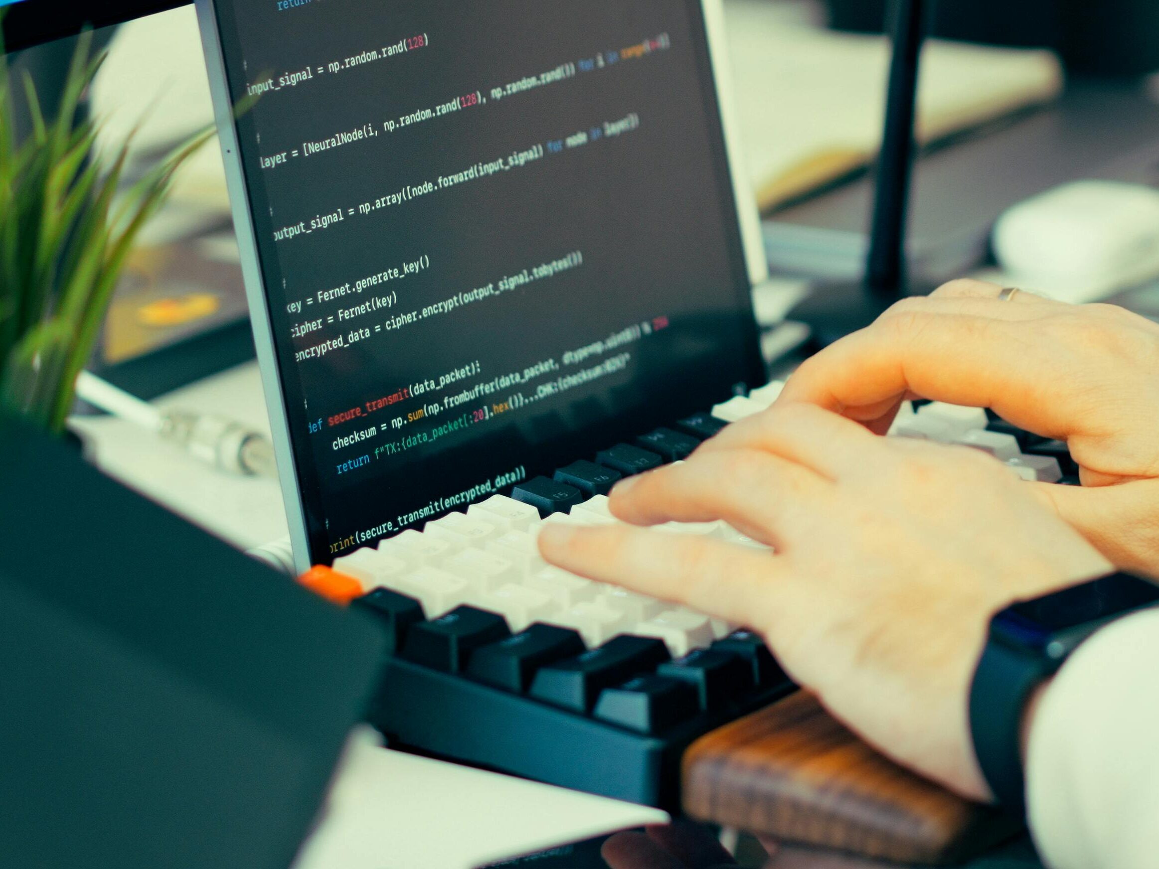 Close-up view of a developer typing code on a keyboard with a computer screen showing scripts.